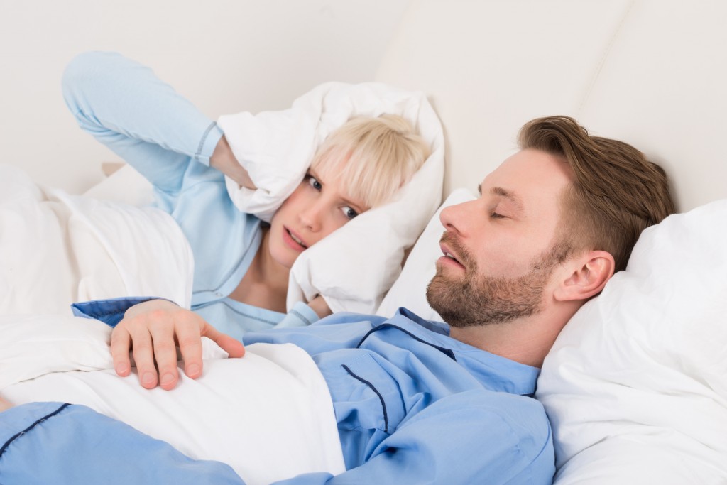 Young Woman Covering Her Ears With Pillow While Man Snoring At Home