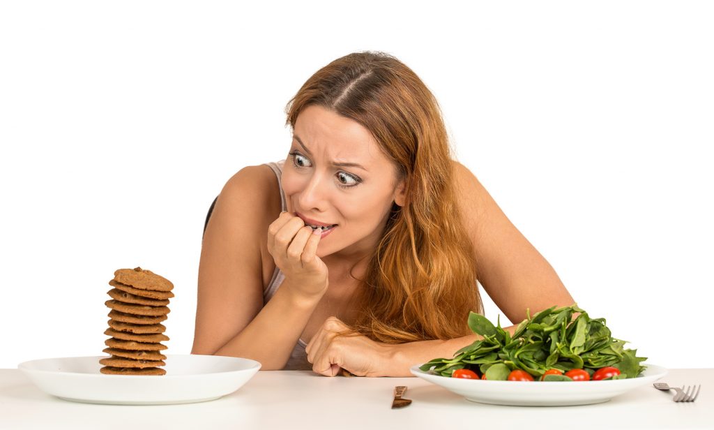 Portrait young woman deciding whether to eat healthy food or sweet cookies she is craving sitting at table isolated white background. Human face expression emotion reaction. Diet nutrition concept